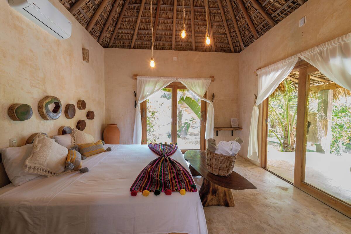 A room with a bed, a window with curtains, a basket, and a thatched roof.