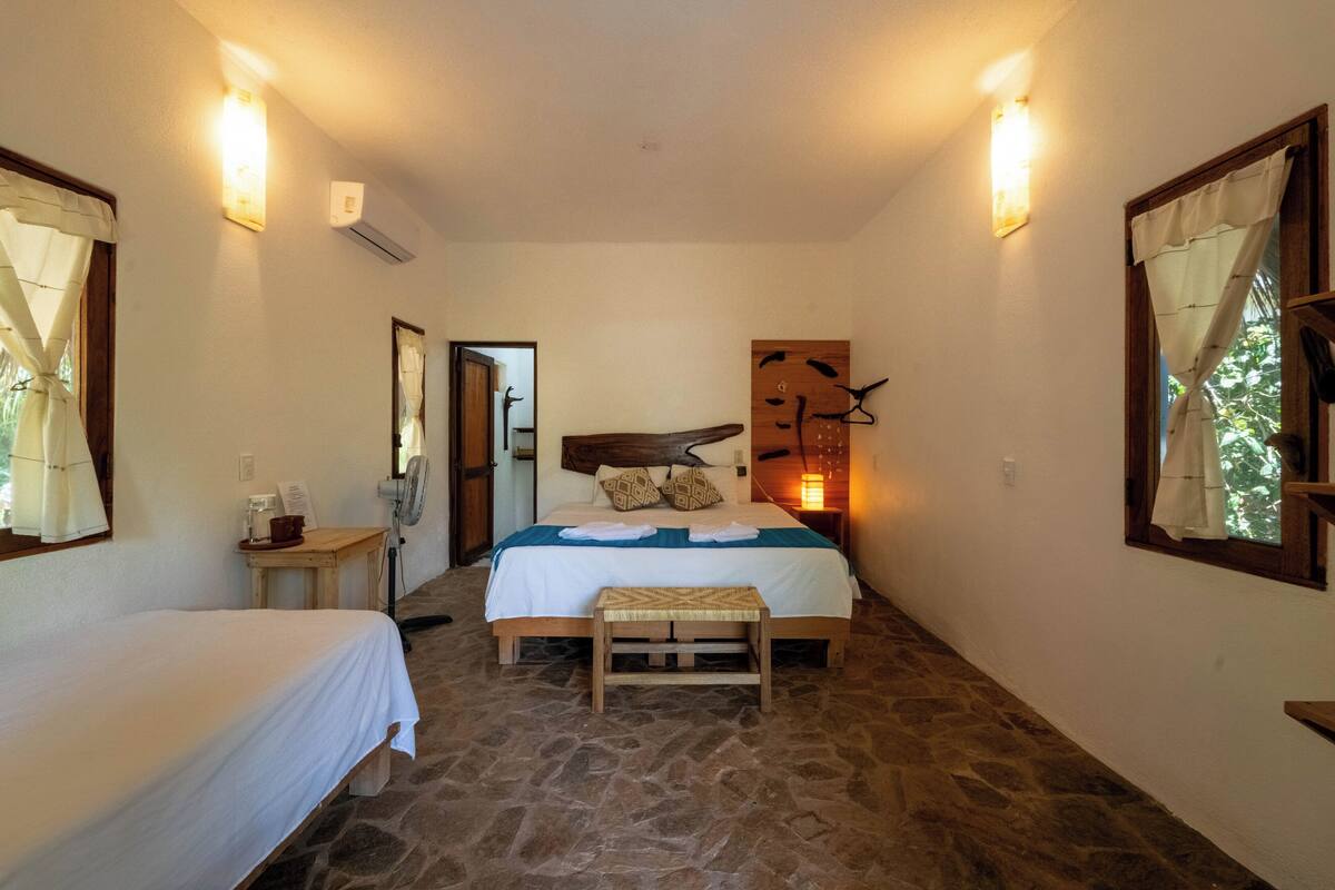A room with a bed, a bench, a small table, and a wall-mounted fan.