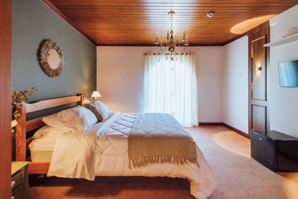 Een slaapkamer met een houten plafond, een bed met witte beddengoed, een nachtkastje met een lamp en een televisie aan de muur.