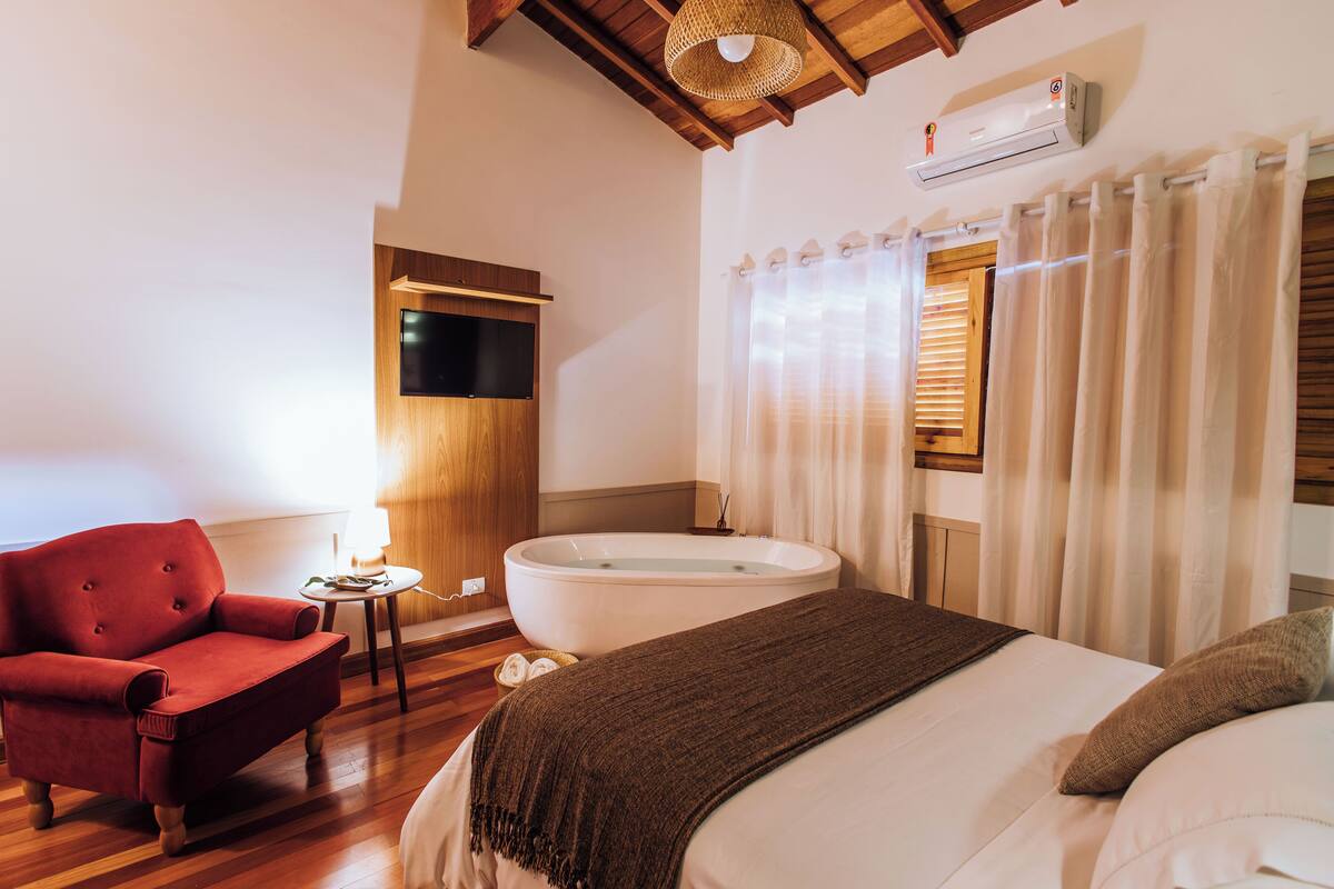 Een slaapkamer met een groot bed, een rode fauteuil, een houten vloer, een plafondventilator en een raam met gordijnen.