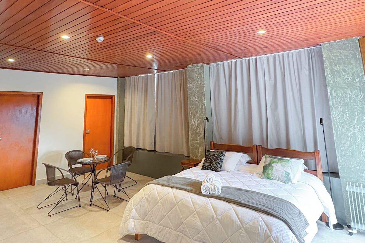 Een slaapkamer met een houten plafond, een bed met een wit sprei, een klein tafeltje met stoelen en een raam met gordijnen.