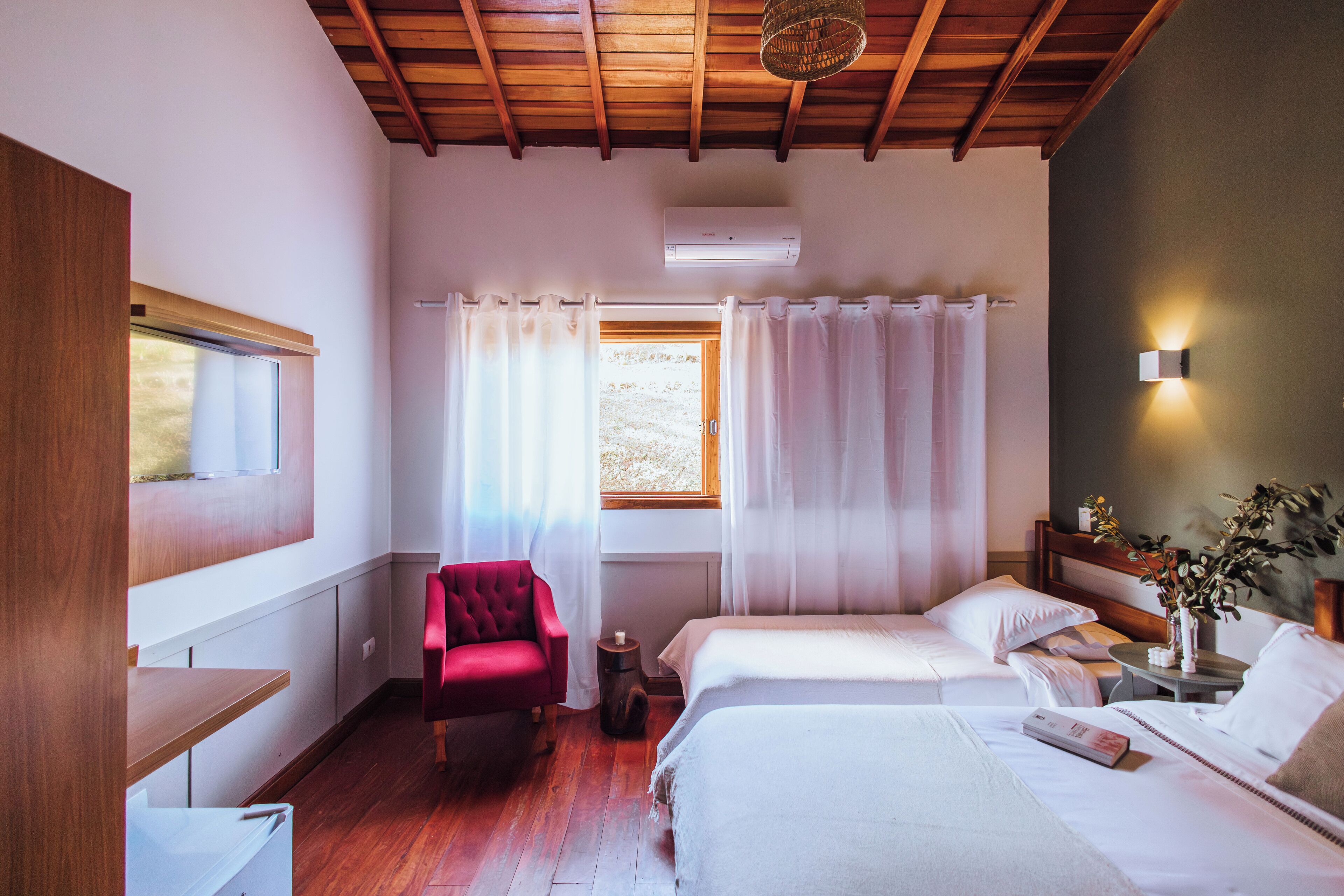 A bedroom with two beds, a red chair, a bedside table with a vase, and a window with curtains.