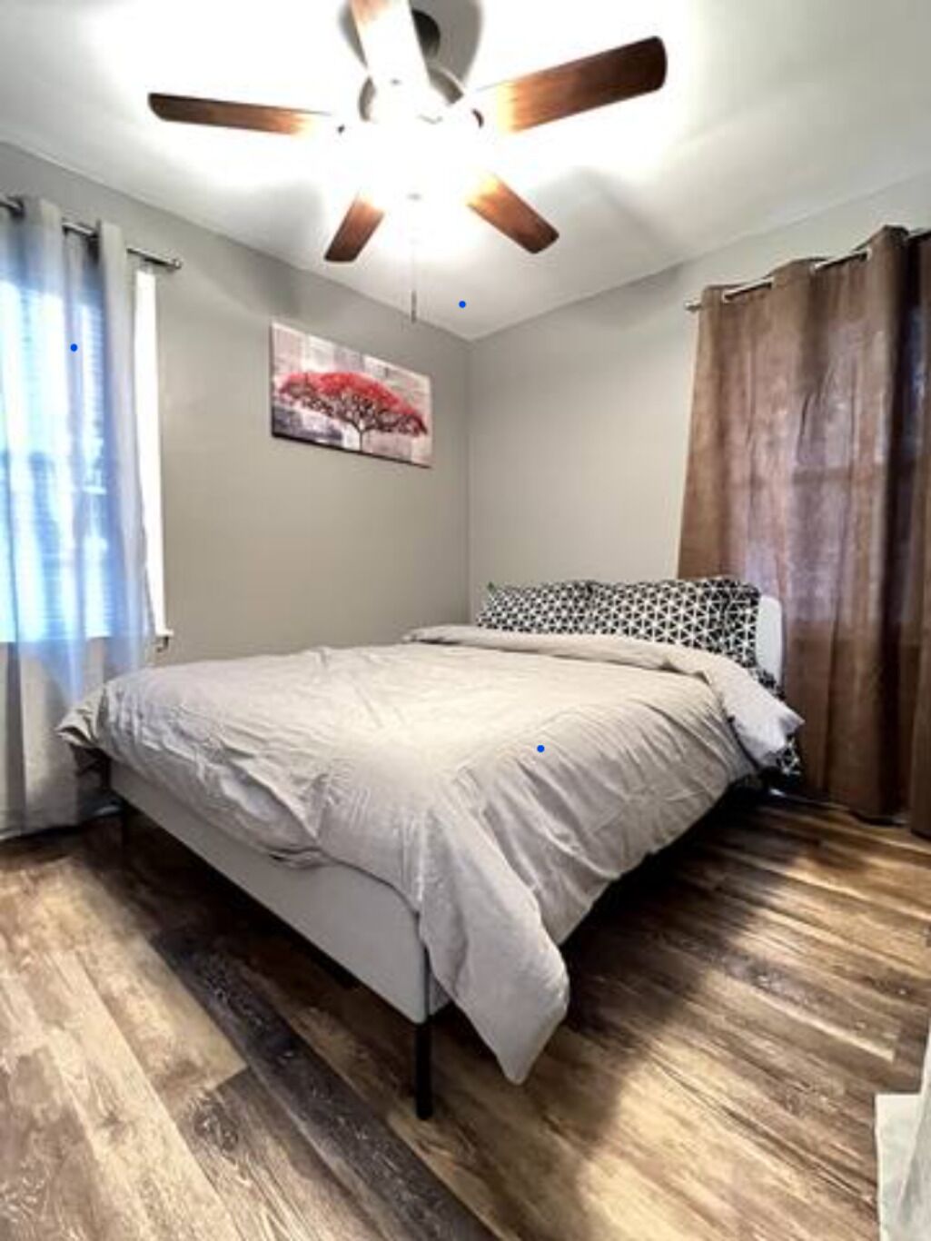 Un dormitorio con una cama, suelo de madera, ventilador de techo, una ventana con cortinas e imagen colgada en la pared.