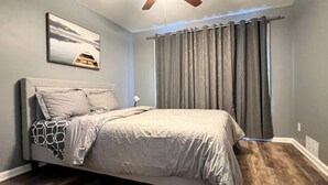 Ein Schlafzimmer mit Bett, Deckenventilator, Fenster mit Vorhängen und einem Bild an der Wand.