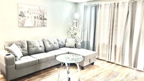 Ein Wohnzimmer mit einem grauen Ecksofa, einem Glastisch, einer weißen Stehlampe und einem großen Fenster mit grauen Vorhängen.