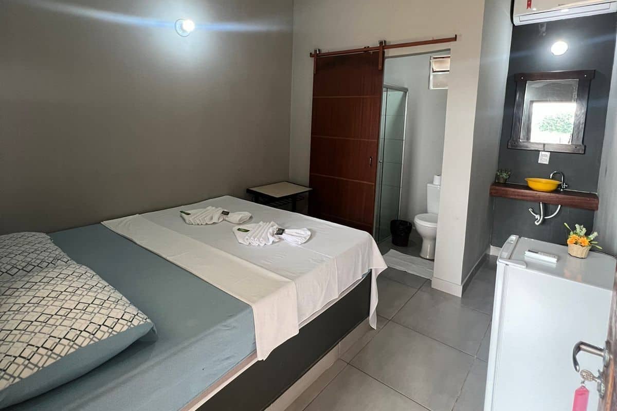 A compact hotel room with a single bed, a small table, a white refrigerator, a bathroom with a toilet, and a sink.