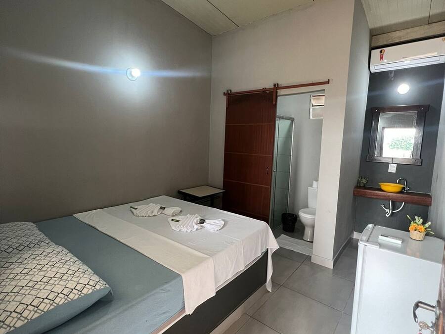 Habitación de hotel pequeña con una cama individual, una mesita, un refrigerador blanco y un baño con inodoro y lavamanos.