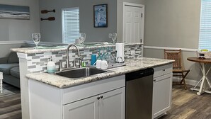 A modern kitchen with a central island, white cabinetry, and a dishwasher. There are pendant lights above the island, a window with blinds, and a dining area with a table and chairs.