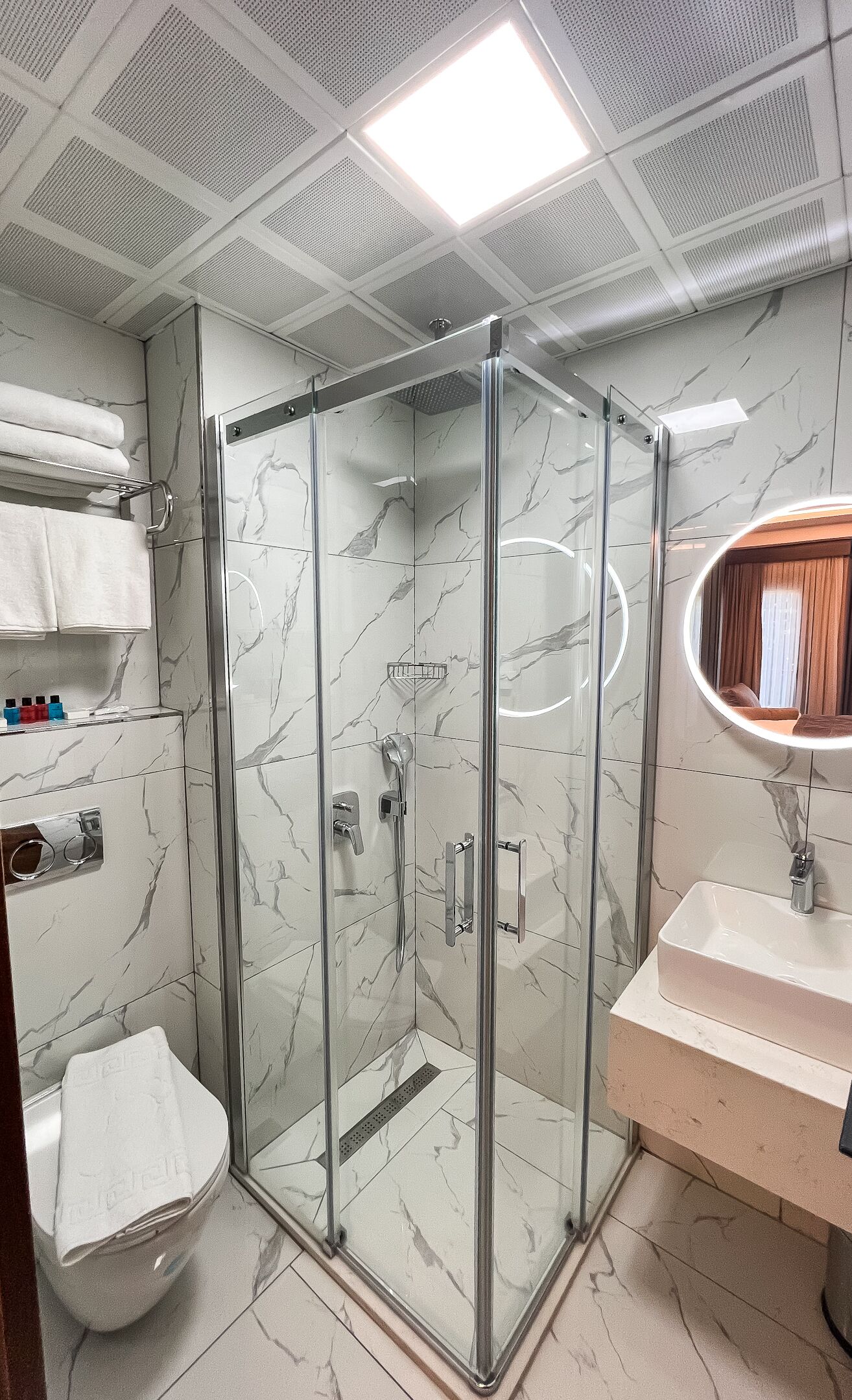 A modern bathroom with a glass-enclosed shower, marble walls, a vanity with a mirror, and a toilet.