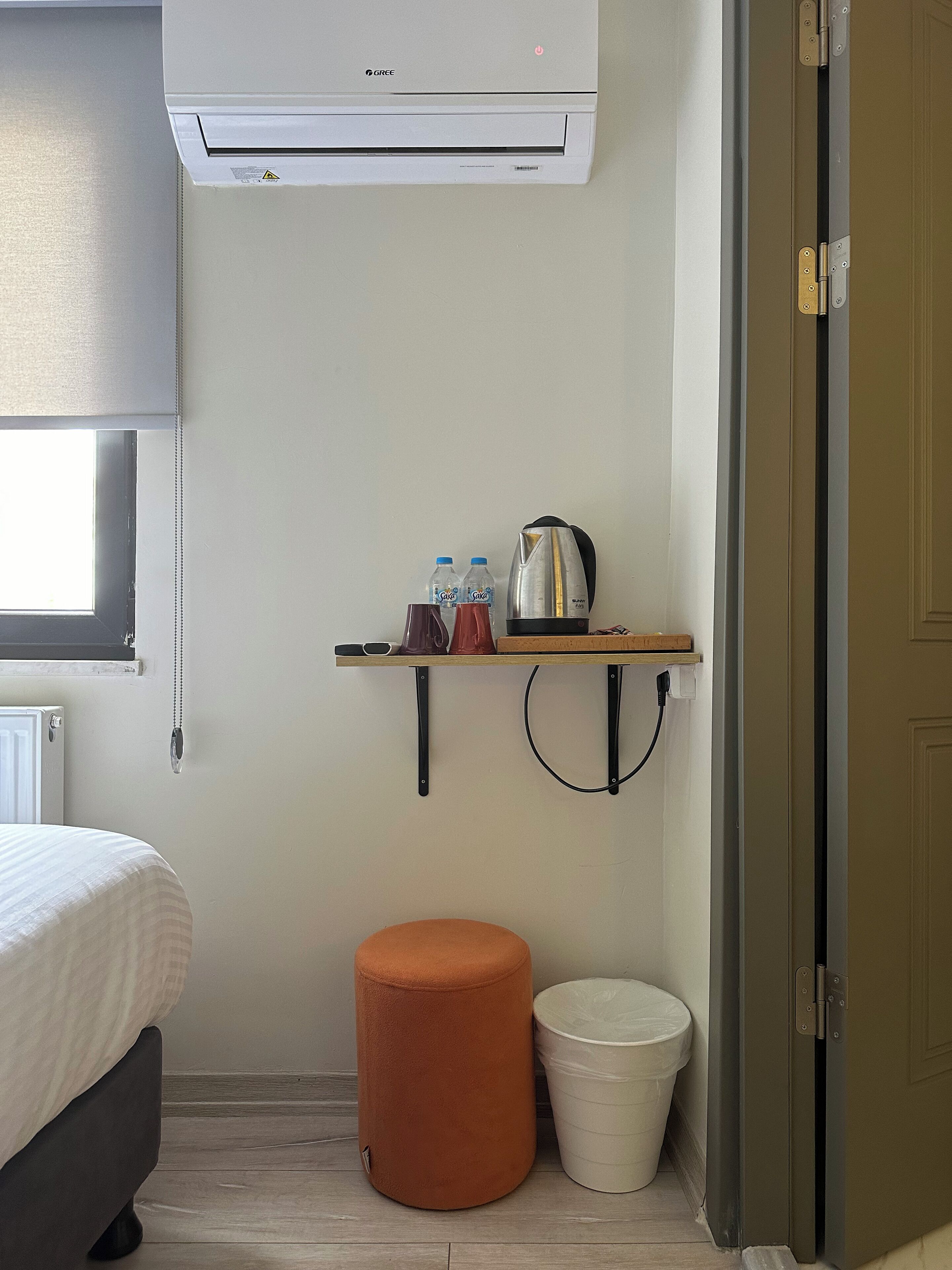 Habitación compacta con cama, aire acondicionado, una pequeña estantería con botellas y tetera, un cubo de basura y una puerta.