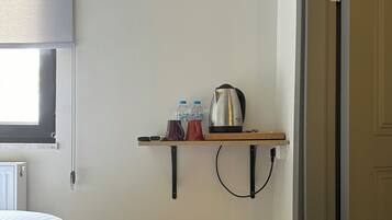 Habitación compacta con cama, aire acondicionado, una pequeña estantería con botellas y tetera, un cubo de basura y una puerta.