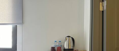 Habitación compacta con cama, aire acondicionado, una pequeña estantería con botellas y tetera, un cubo de basura y una puerta.