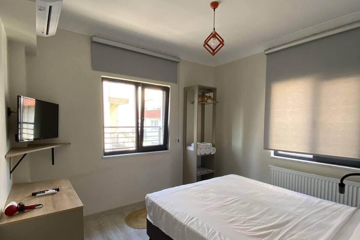A bedroom with a bed, a desk, a chair, a radiator, a window, and a hanging light fixture.