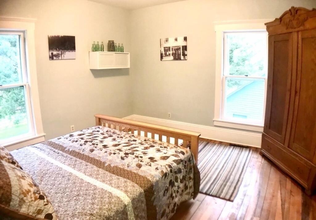A bedroom with a bed, a wooden bench, a small shelf with bottles, and two windows.