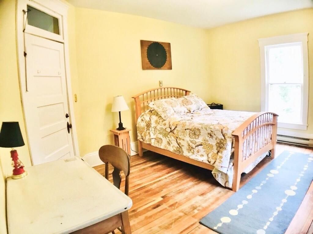A bedroom with a wooden bed, a desk with a lamp, a chair, and a window with curtains.