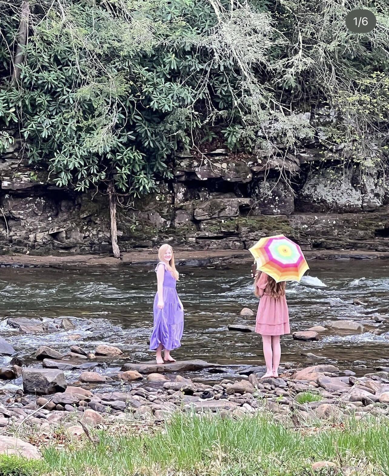 Two people standing by a rocky riverbank with trees in the background.