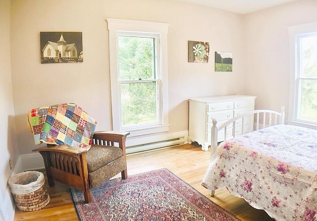 A bedroom with a wooden chair, a white dresser, a bed with a floral bedspread, a window, and a rug.
