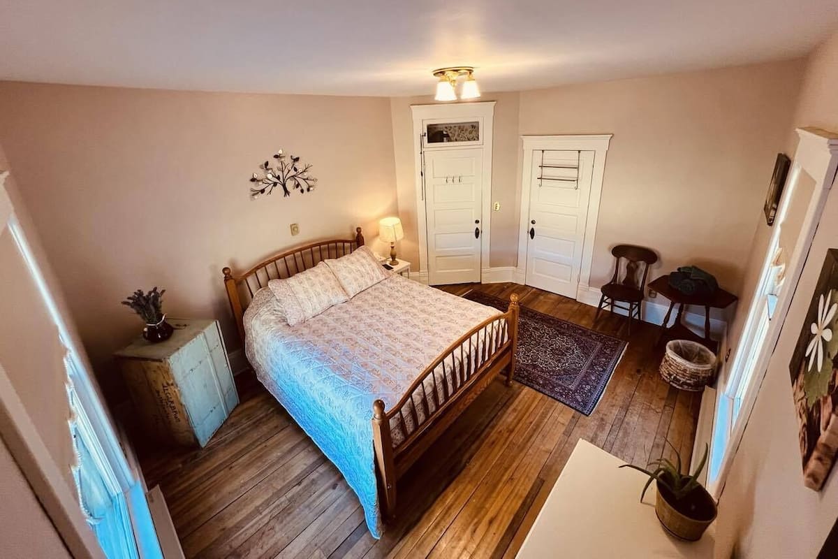 A bedroom with a wooden bed, a nightstand, a lamp, a door, a small table, and a chair.