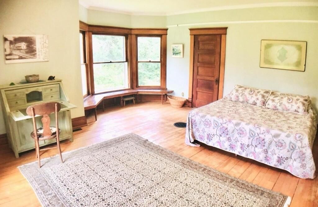 Un dormitorio con cama, escritorio, silla, asiento junto a la ventana y un ventanal grande.