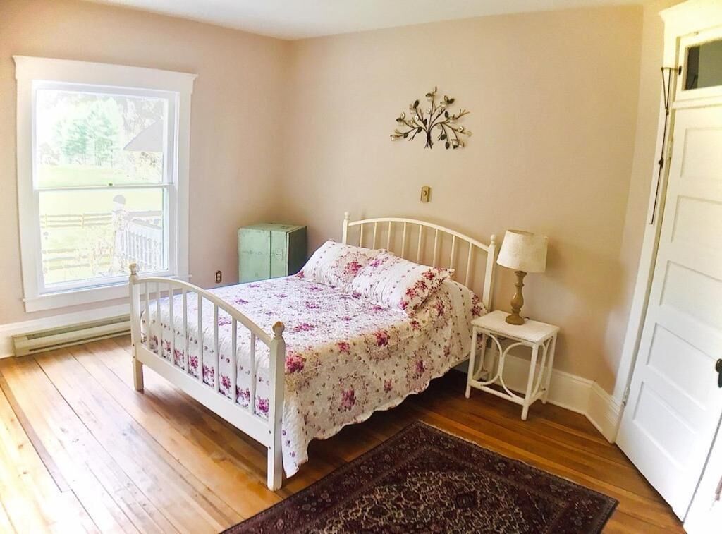 A bedroom with a wooden floor, a white bed with a floral bedspread, a nightstand with a lamp, and a decorative wall piece.