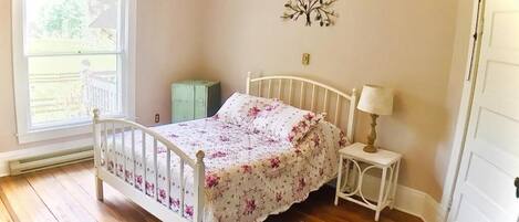 A bedroom with a wooden floor, a white bed with a floral bedspread, a nightstand with a lamp, and a decorative wall piece.