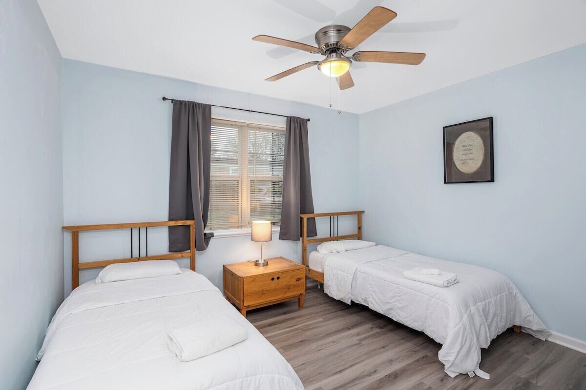 A bedroom with two single beds, a ceiling fan, a bedside table with a lamp, and a window with curtains.