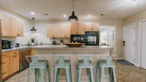 A modern kitchen with wooden cabinets, a central island with stools, a black oven, and a hanging pendant light. - 4BR/3BA w/POOL in golf cart district near the Beach - Shadowlawn (Ocean Springs)
