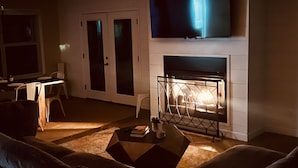A dimly lit living room with a fireplace, a flat-screen TV mounted above it, and a sofa in the foreground. - 4BR/3BA w/POOL in golf cart district near the Beach - Shadowlawn (Ocean Springs)
