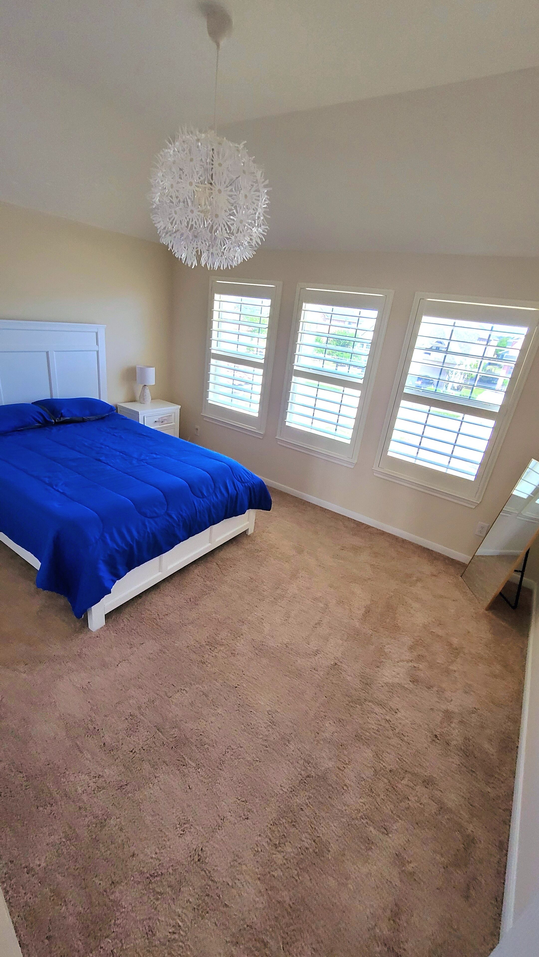 A bedroom with a large bed, a white nightstand, a white lamp, and three windows with blinds.