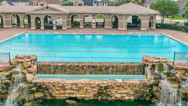 A swimming pool with a stone waterfall feature, surrounded by a building with arched walkways and an American flag. -  truly stunning lakefront with immediate access to all the city has to offer. (Cypress)