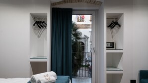 Une chambre d’hôtel avec un lit, une porte-fenêtre donnant sur un balcon, un porte-manteau mural et une petite étagère avec une radio.