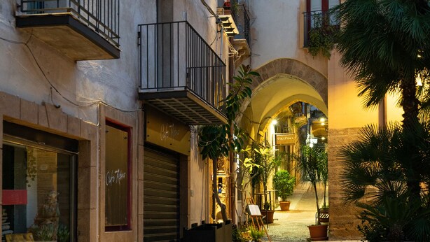 Une ruelle urbaine bordée d’immeubles, agrémentée de plantes en pot et surmontée d’une petite arche.