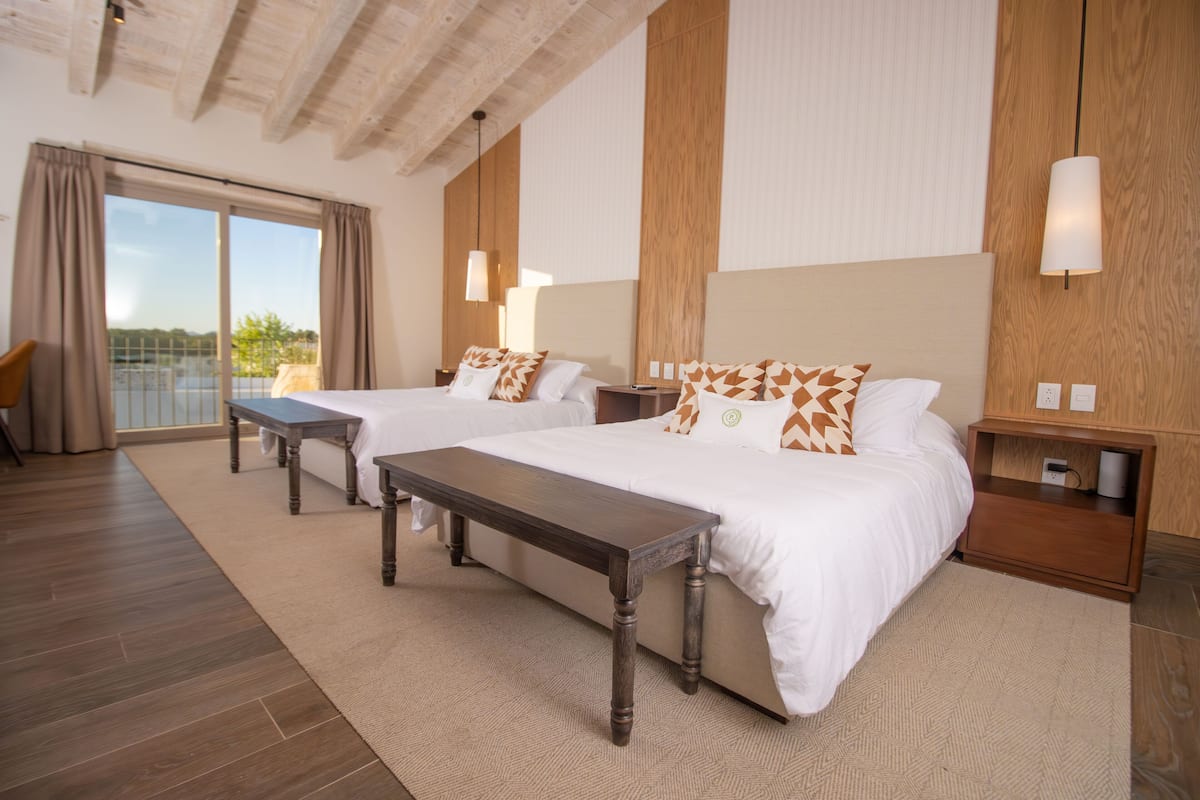 A hotel room with two beds, a wooden bench, a large window with curtains, and a wooden nightstand with a lamp.