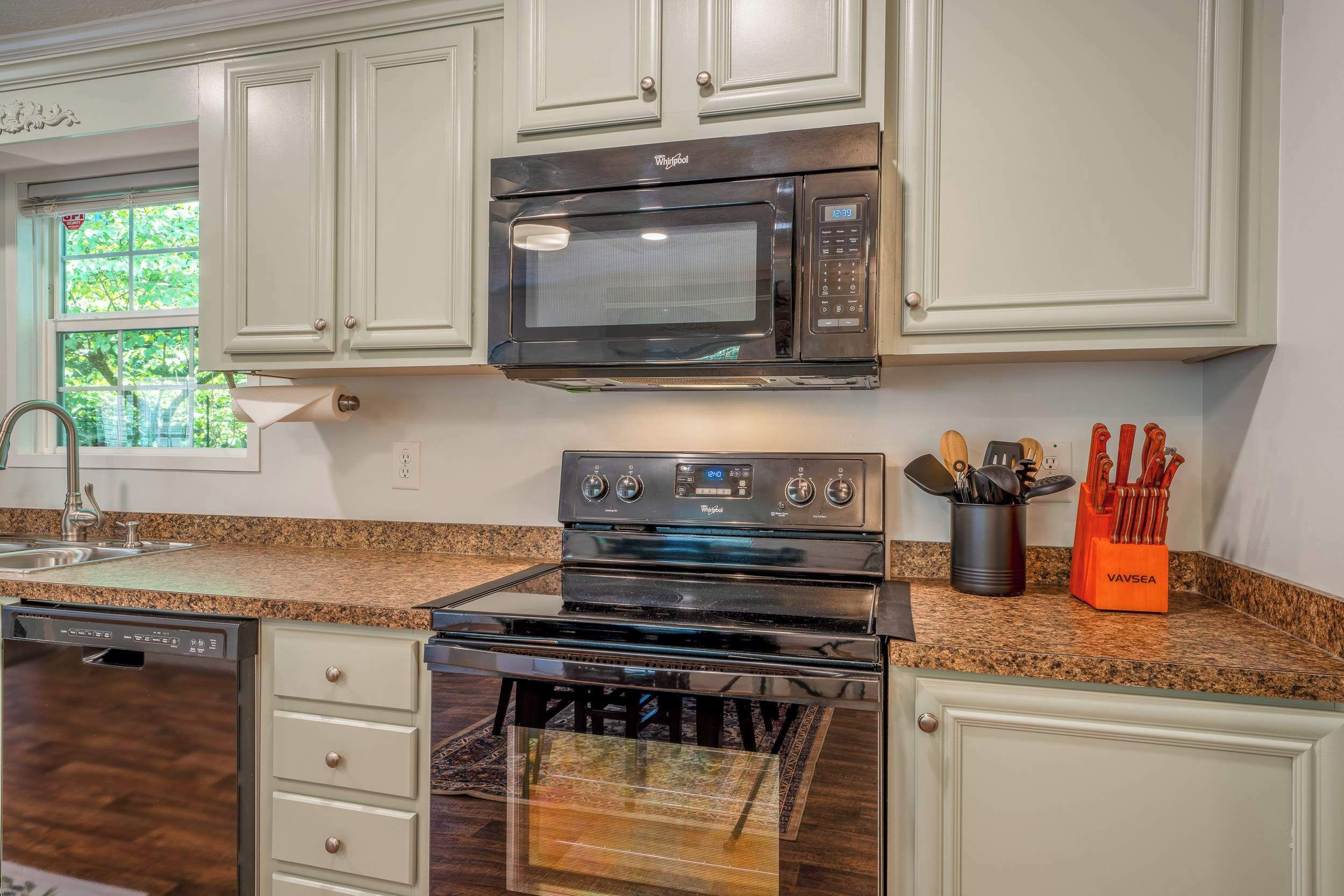A kitchen with white cabinetry, a stainless steel microwave, a double oven, and a dishwasher. There is a window with a view of greenery outside.