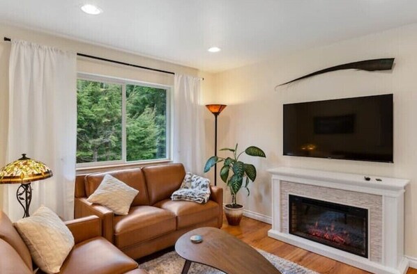 A living room with a leather sofa, a fireplace, a television, and a large window with curtains. - Cozy 2BR Apt, Hot tub, King bed, secluded, and freshly remodeled!
 (Juneau)
