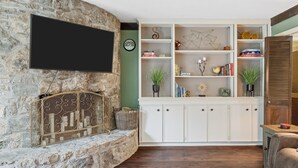 Un salon avec un mur en pierre, un téléviseur à écran plat, un meuble blanc avec des étagères et une petite table sur laquelle est posée une plante.