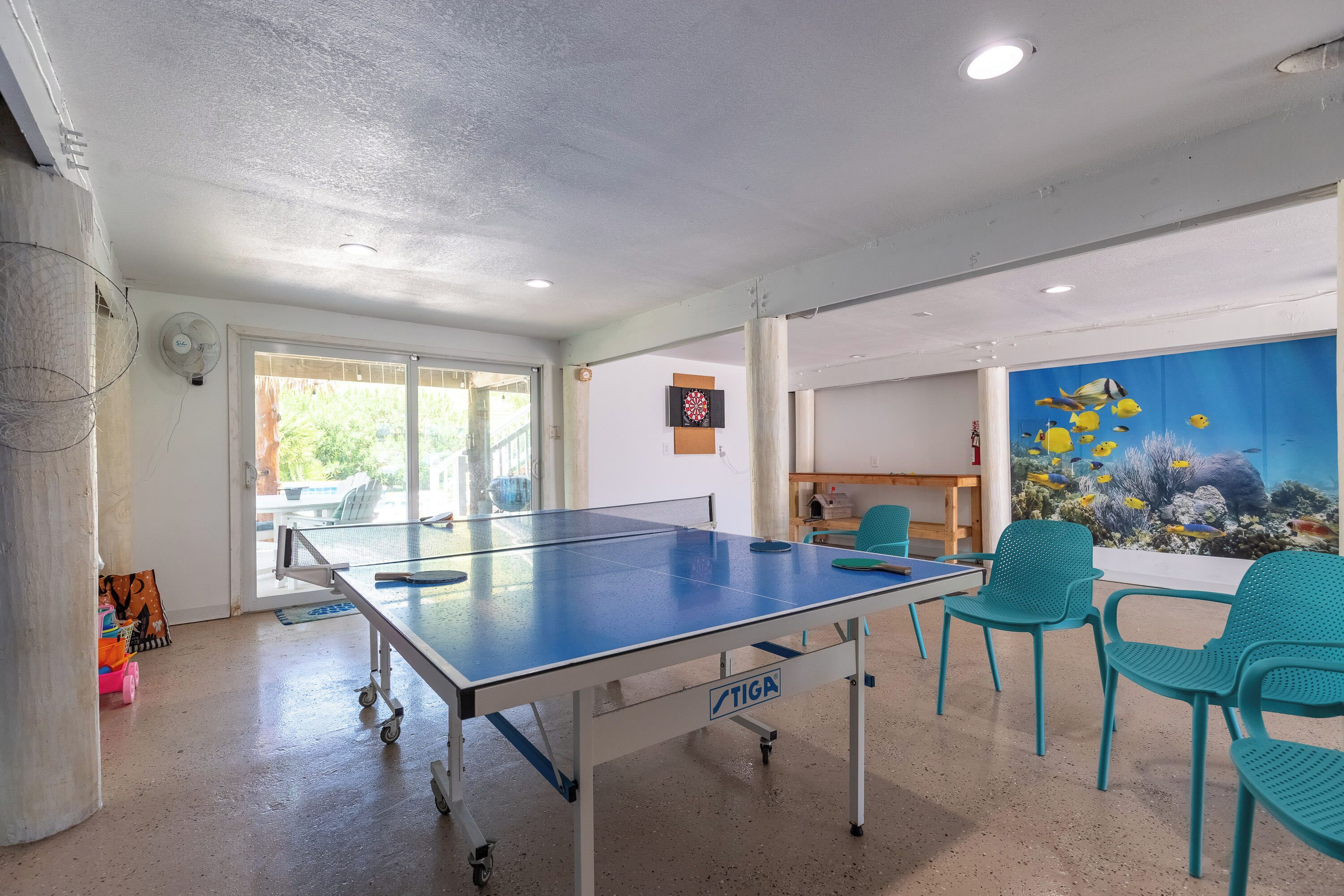 Um quarto com mesa de pingue-pongue, cadeiras verde-água, alvo de dardos e um grande mural com tema de peixes.