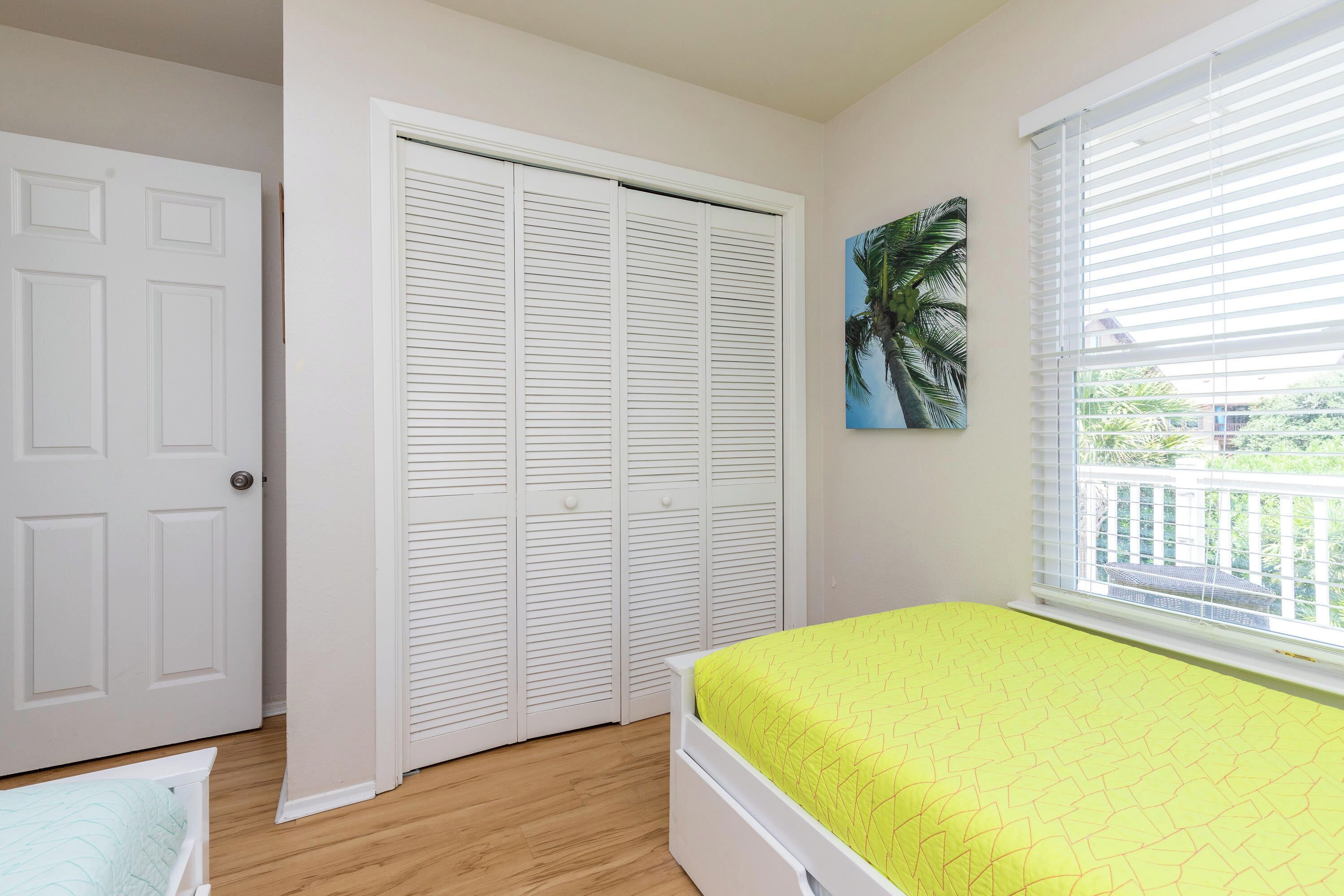 A bedroom with a bed, a closet, a window with blinds, and a painting of a palm tree.
