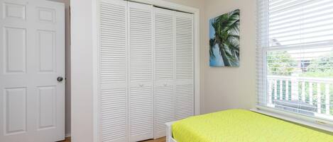 A bedroom with a bed, a closet, a window with blinds, and a painting of a palm tree.