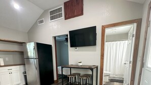 Eine moderne Küche mit Kühlschrank, Holzschränken, einem Esstisch mit Hocker, einem an der Wand montierten Fernseher und einem Deckenventilator.