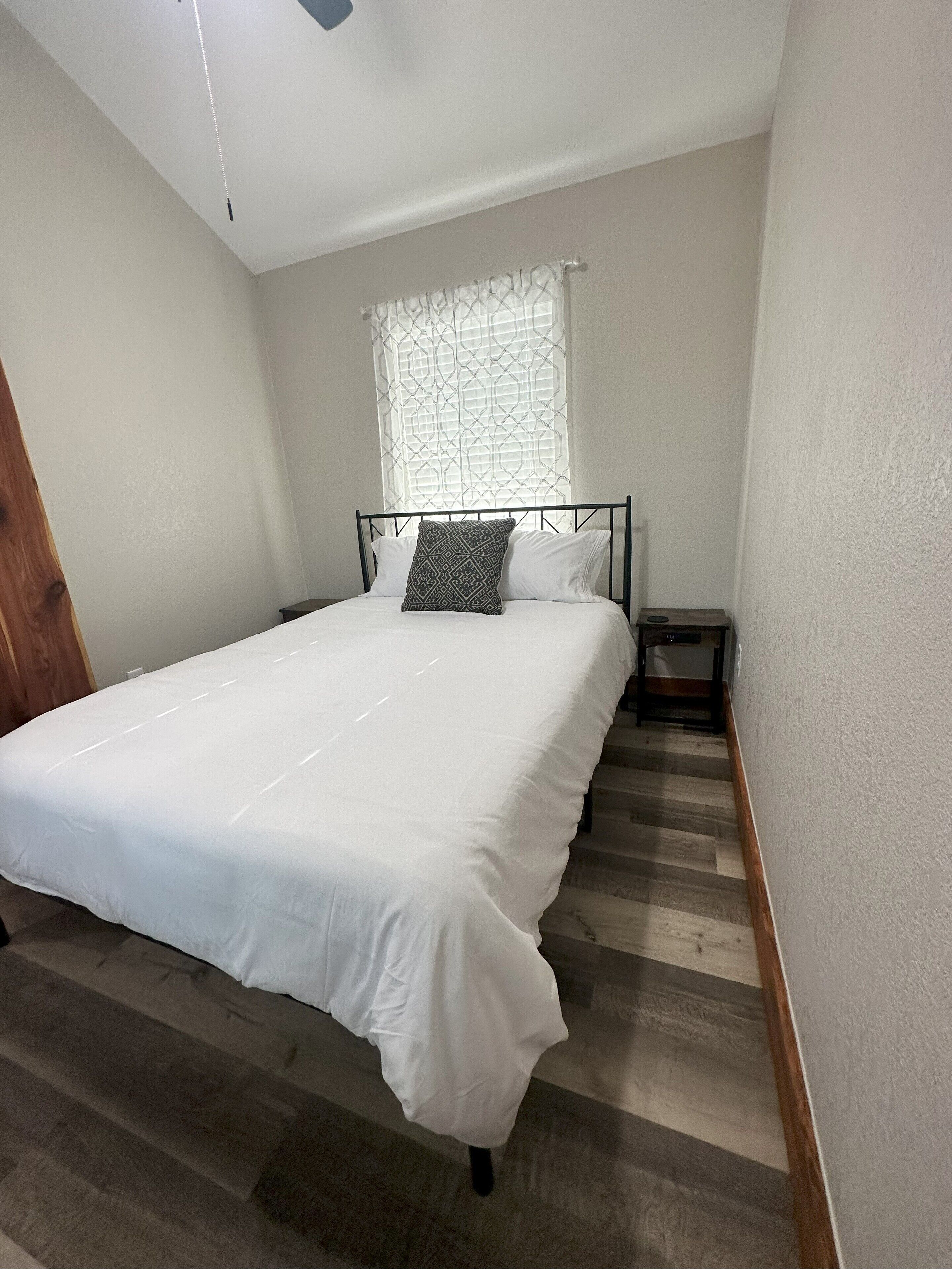 Un dormitorio con una cama, una mesita de noche, un ventanal con cortina estampada y una puerta de madera.