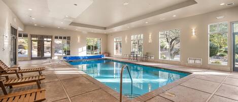 An indoor swimming pool with clear glass walls, wooden lounge chairs, and a blue deck area.