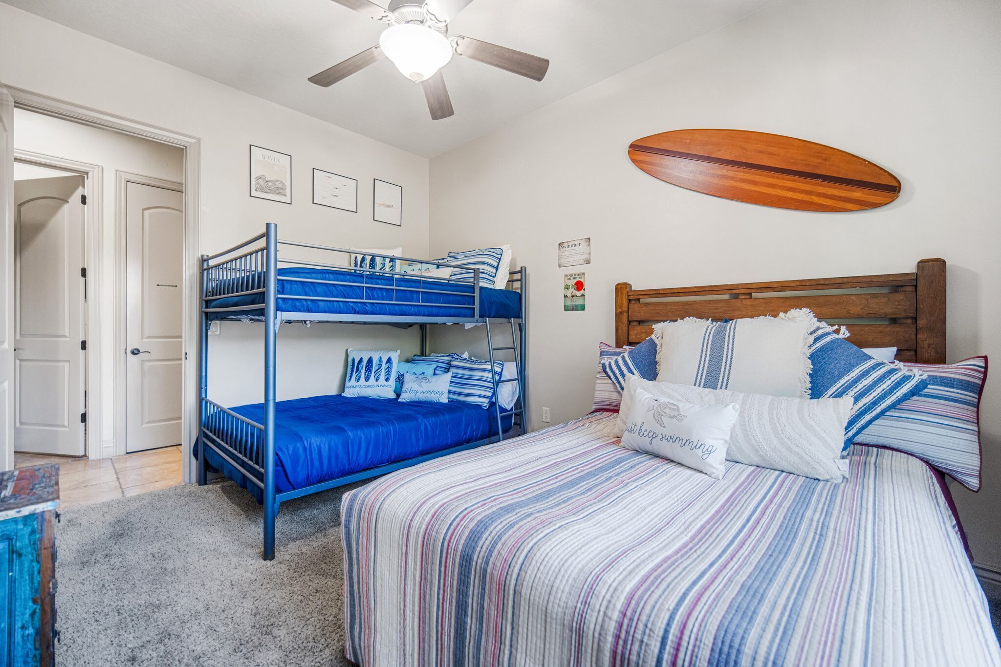 Une chambre avec des lits superposés, un lit simple, un ventilateur de plafond et une planche de surf accrochée au mur.