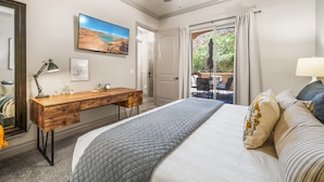 Un dormitorio con una cama, un escritorio de madera, un ventilador de techo, un cuadro y vistas a través de una ventana.