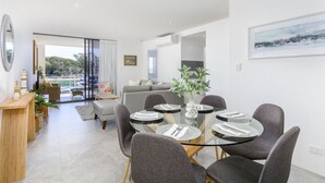 Un área de comedor moderna con una mesa de vidrio y sillas grises tapizadas, un sofá gris y vistas al exterior.