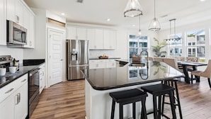 A modern kitchen with white cabinetry, stainless steel appliances, a central island with bar stools, and a dining area with a view of the outside.