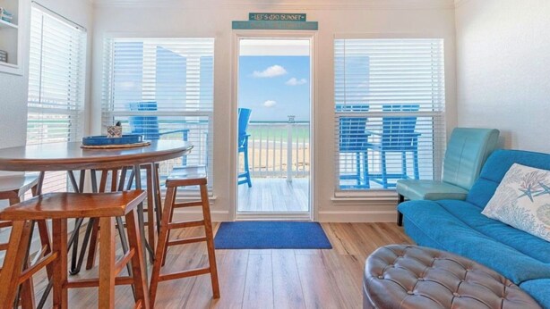 A room with a wooden dining table, blue chairs, a blue sofa, and a view of the beach through a door. - GulfScape2217 Seascape Condos 1bed1bath Beachfront Condo Ocean View from balcony (Galveston)