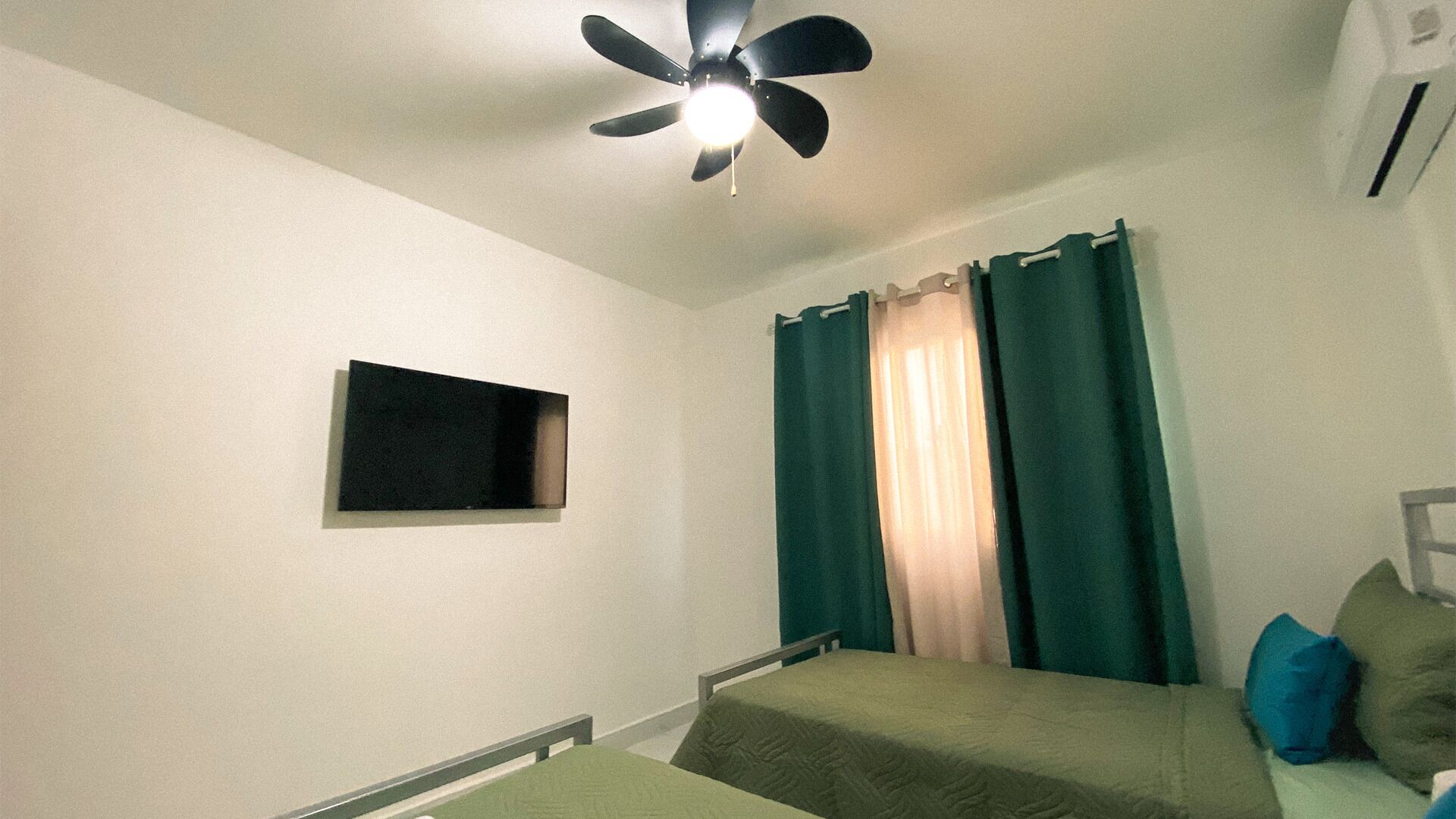 A small, simple room with a bed, a ceiling fan, a window with curtains, and a wall-mounted air conditioning unit.