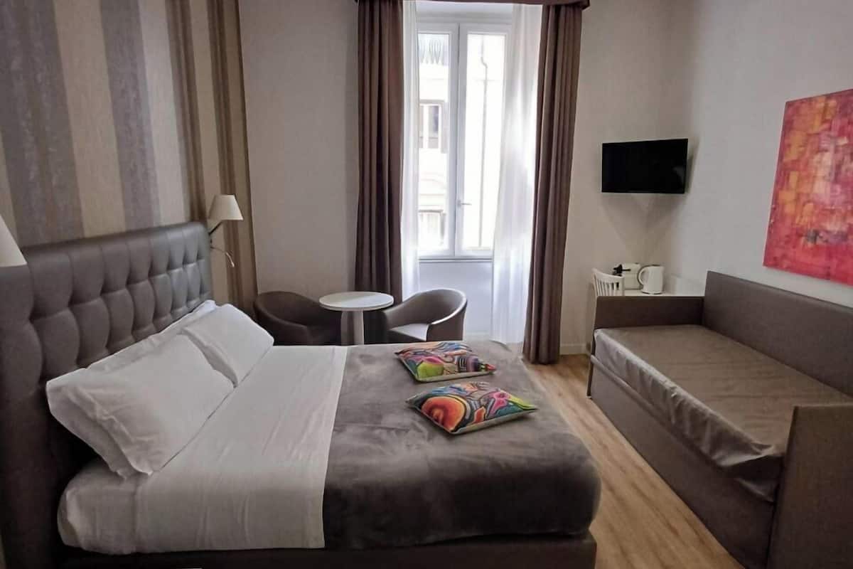 Habitación de hotel con una cama grande, un sofá, una mesita, un televisor y una ventana con cortinas.