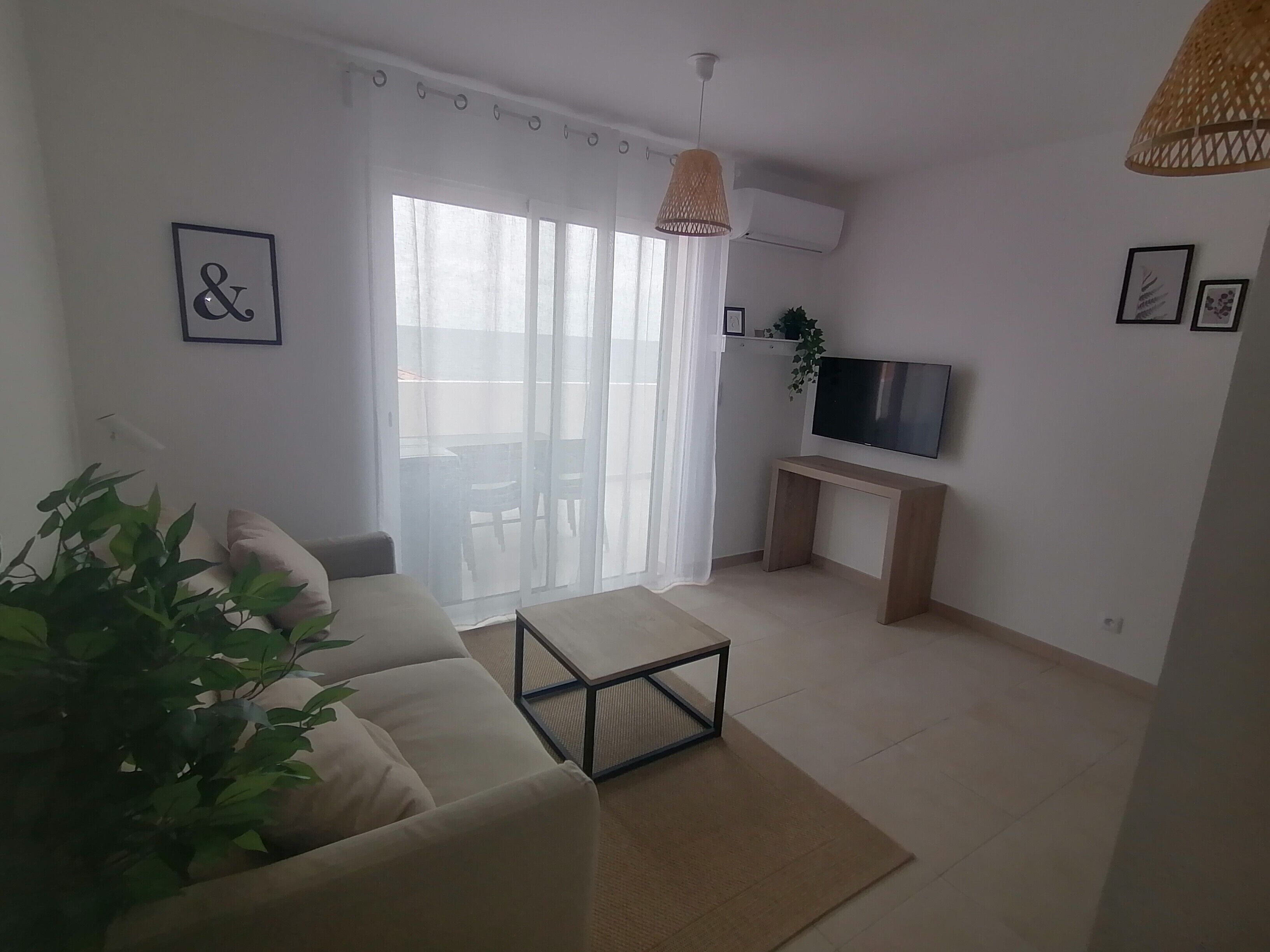 Un salon comprenant un canapé, une table basse, un téléviseur posé sur un meuble et une porte-fenêtre coulissante donnant sur un balcon.
